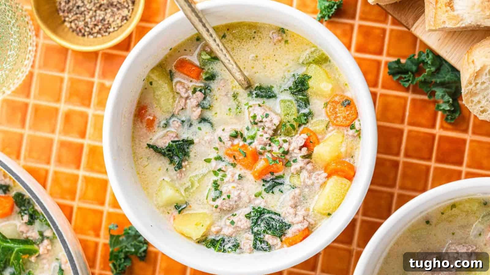 A bowl of creamy soup with sausage, kale, carrots, and potatoes, with a spoon inside, on an orange tiled surface.
