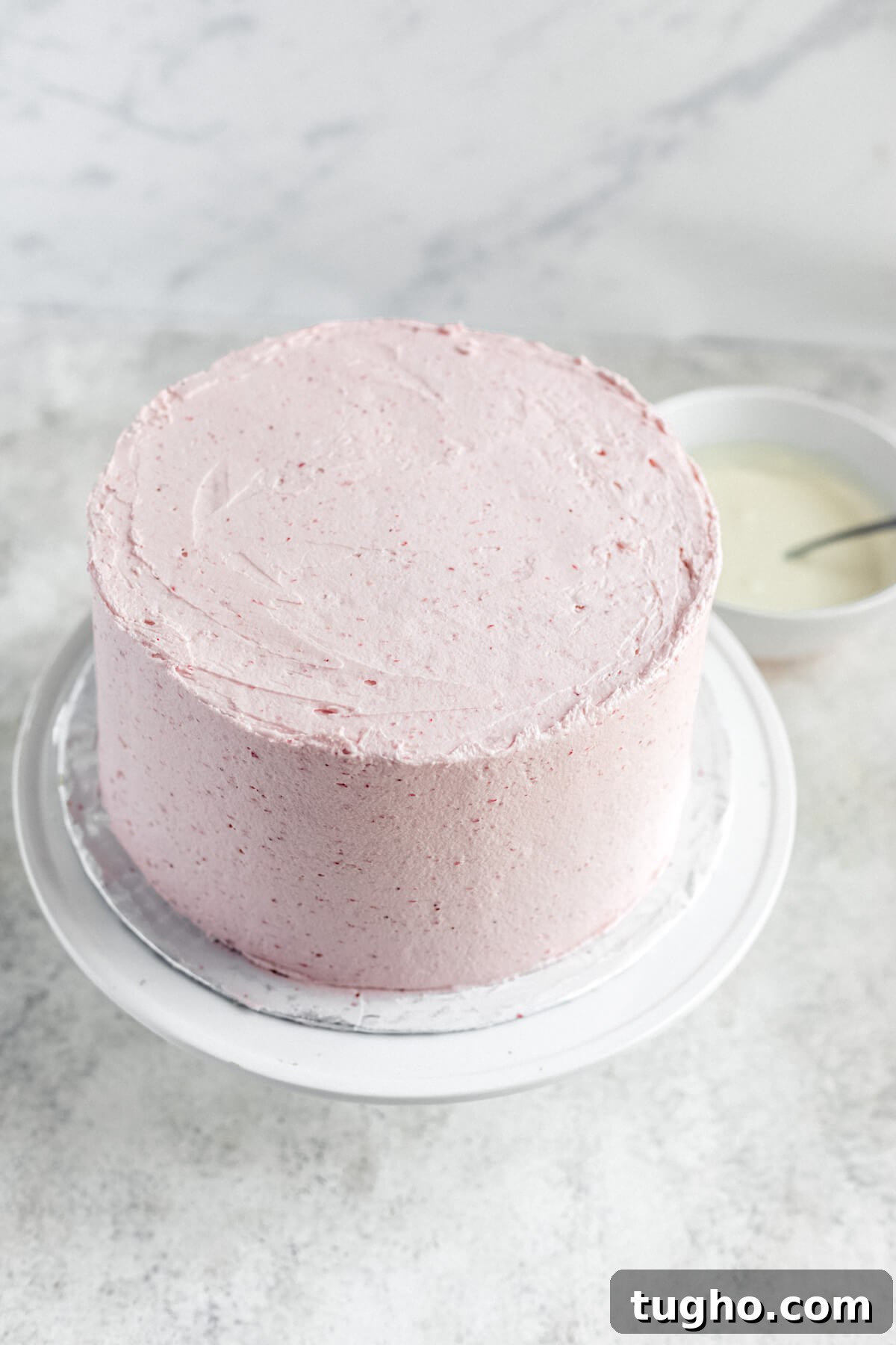 White Chocolate-Covered Strawberry Cake Recipe 8 frosted cake on a cake stand with white chocolate ganache in the background