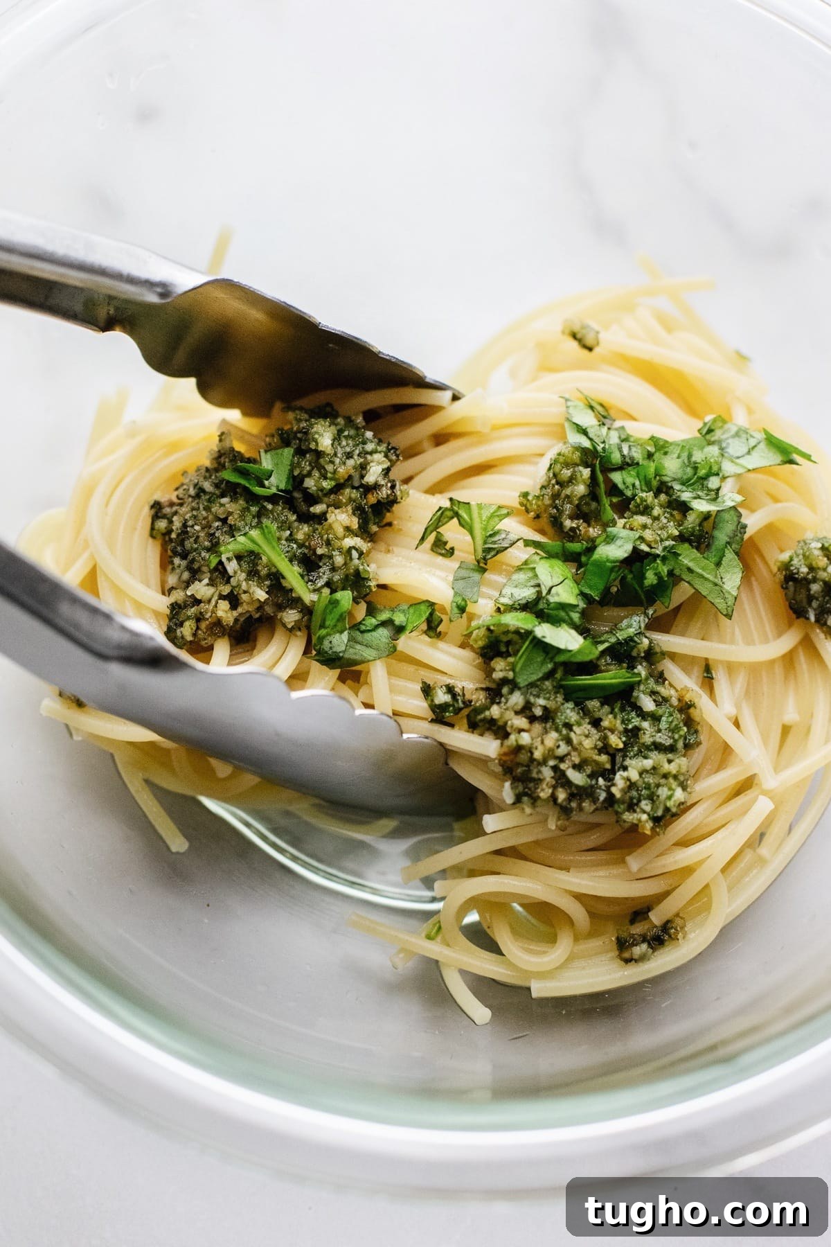 Pesto Spaghetti with Fresh Garden Tomatoes 4 Tongs tossing spaghetti and pesto in a clear glass bowl.