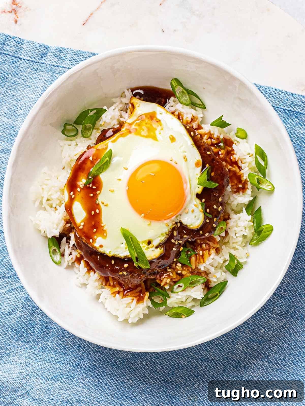 Loco Moco Recipe: Hawaiian Burger with Savory Brown Gravy 9 Hawaiian loco moco in a white bowl with a beef burger patty, brown gravy, and a sunny side up egg on top of a blue napkin.
