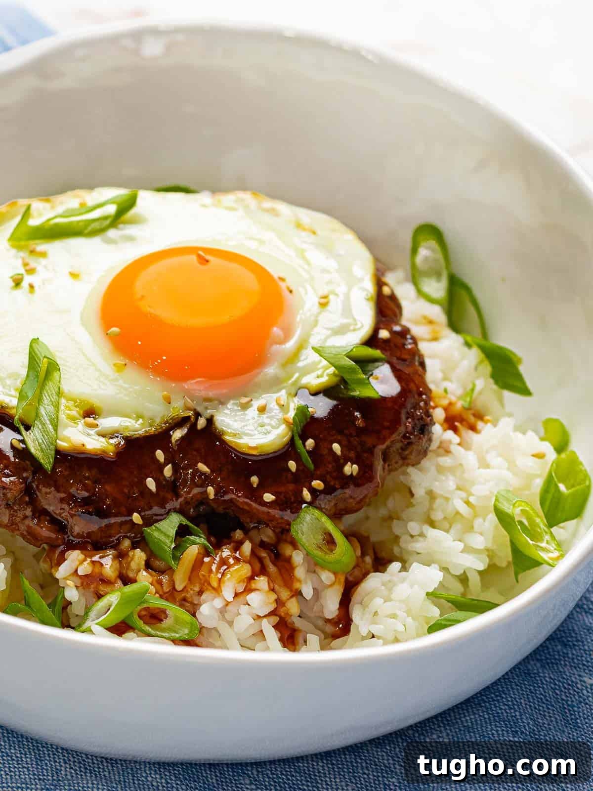 Loco Moco Recipe: Hawaiian Burger with Savory Brown Gravy 3 Loco moco in a white bowl filled with white rice, beef burger, brown gravy, and a fried egg that is garnished with green onions.